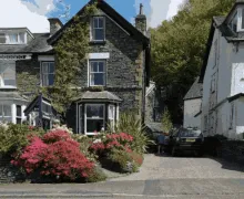 An external view of the three-star  Brooklands - Guest house near Bowness-on-Windermere, a traditional lakeland stone built property with mature planting in the front garden and parking.