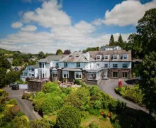 A view of the large Windermere Hillthwaite Hotel and gardens panted with small trees and shrubs in rolling countryside.